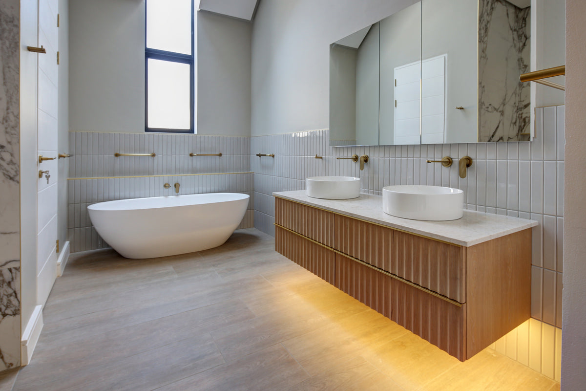 A modern bathroom interior with a freestanding white bathtub, double vessel sinks on a wood-look vanity with a marble countertop, large mirrors, and warm under-vanity lighting.