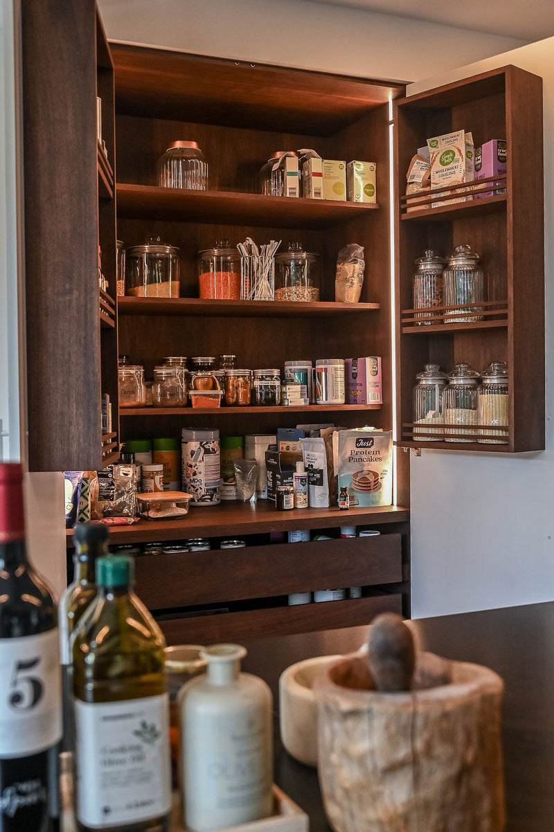 A tall dark wood pantry cabinet with its doors open, filled with glass jars, boxes, and bottles of pantry items. The cabinet is set in a kitchen environment with a countertop in the foreground containing bottles and a wooden mortar and pestle.