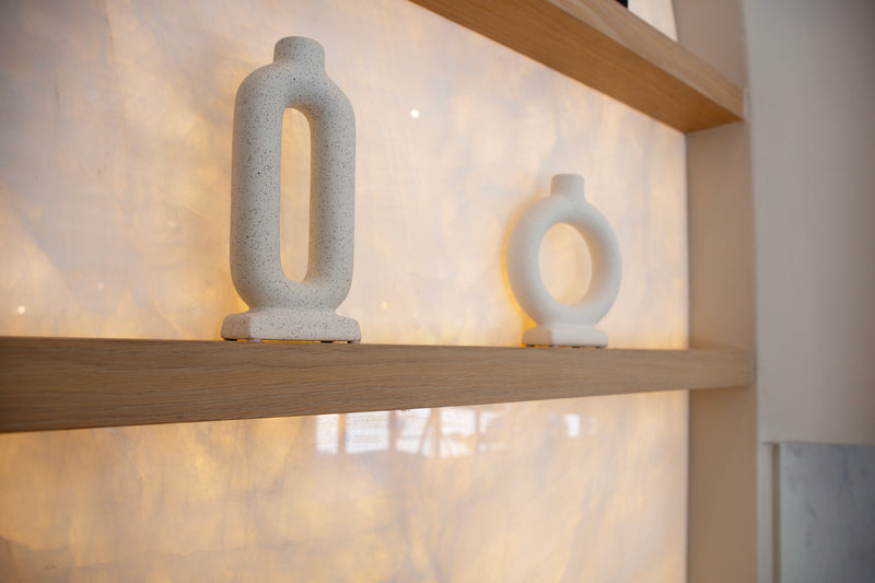 Two modern minimalist stone vases in a light speckled white finish, displayed on a wooden shelf against a warm, illuminated marble wall.