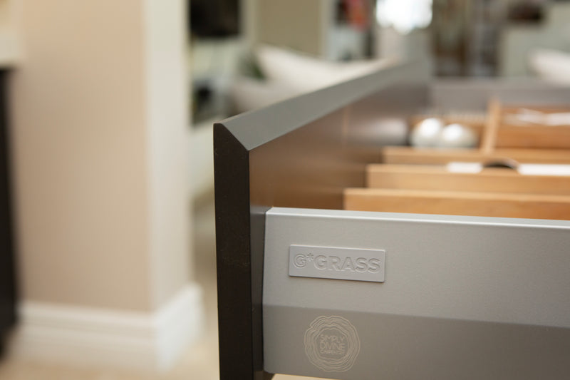 Close-up of a modern kitchen drawer with a silver front panel, a visible GRASS logo, and a dark front rail, shown in a softly blurred interior setting.
