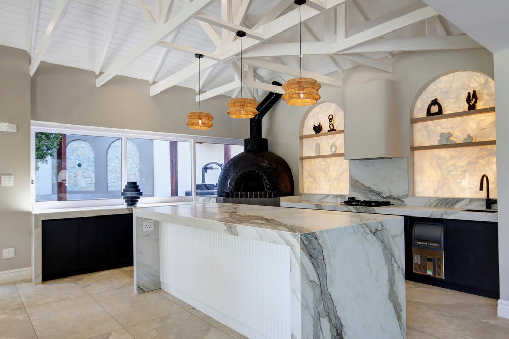 A modern kitchen interior with a large white marble island, black cabinetry, arched built-in shelves with warm lighting, and a dark freestanding wood-burning stove. The room has a high white-beamed ceiling and large windows.